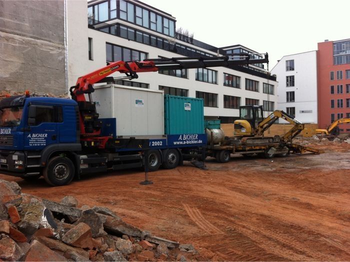 LKW mit Kran hebt Container auf Baustelle mit Gebäuden im Hintergrund