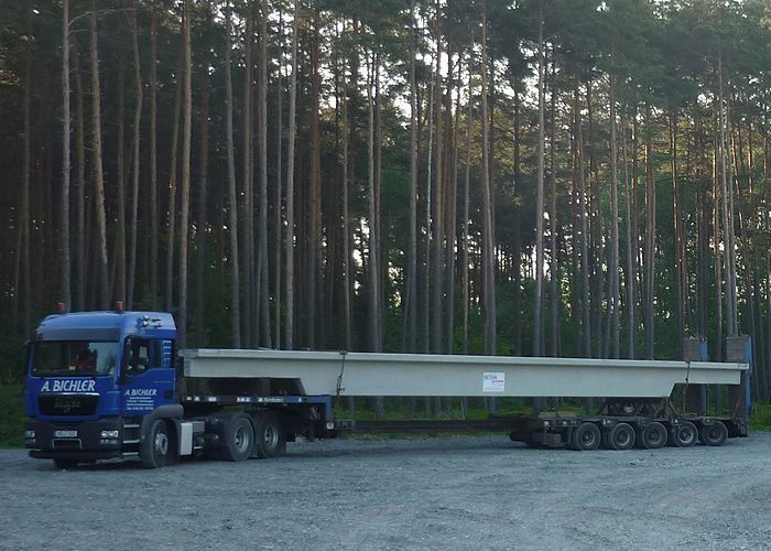 Langer LKW mit Betonfertigteil auf Ladefläche, im Hintergrund Wald