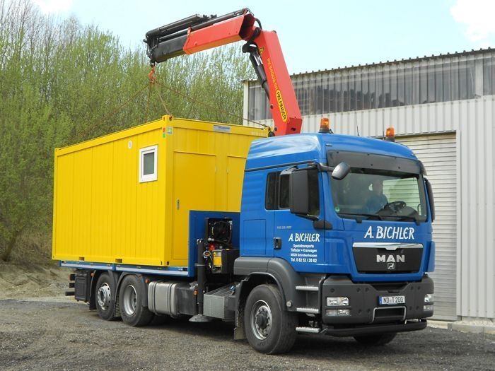 Blauer LKW mit Kran hebt gelben Container an Baustelle ab